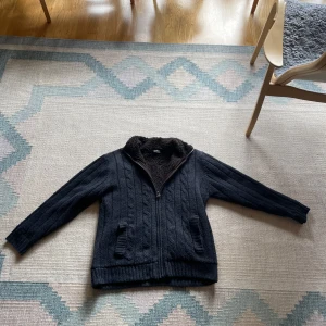 Stickad jacka  - Mysig svart stickad jacka med dragkedja och brunt fuskpälsfoder. Jackan har kabelstickat mönster, hög krage och två fickor framtill. Inga skador eller skavanker. 