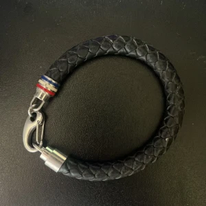 Svart flätat armband med metallspänne från hilfiger - Stilrent svart armband i flätat material med ett robust metallspänne. Detaljer i silverfärgad metall och små ränder i rött, vitt och blått vid låset. Perfekt accessoar för en modern look. Ny pris 749