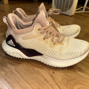 Beige Adidas Bounce sneakers - Säljer ett par snygga Adidas Bounce sneakers i beige med vita och burgundy detaljer. Skorna har en vågig sula och är tillverkade i syntetmaterial med mesh för extra andningsförmåga. Perfekta för dig som gillar sportig stil.