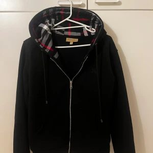 Svart hoodie från Burberry med rutigt foder - Säljer en svart hoodie från Burberry med dragkedja och klassiskt rutigt foder i huvan och insidan. Tröjan har långa ärmar, fickor framtill och justerbar huva med snören. Perfekt för dig som gillar stilrena detaljer och ikoniskt mönster.