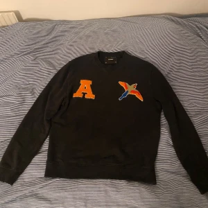 Svart sweatshirt från Axel Arigato med broderad bokstav och fågel - Cool svart sweatshirt från Axel Arigato med en stor orange broderad bokstav 'A' och en färgglad broderad fågel på bröstet. Tröjan har rund hals och långa ärmar. Perfekt för en trendig och avslappnad stil.