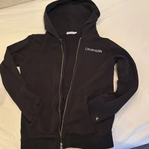 Svart hoodie från Calvin Klein Jeans - Svart hoodie från Calvin Klein Jeans med dragkedja och huva. Logga på bröstet och litet tryck på ärmen samt 'JEANS' patch bak. Perfekt för en avslappnad stil och enkel att matcha med det mesta.