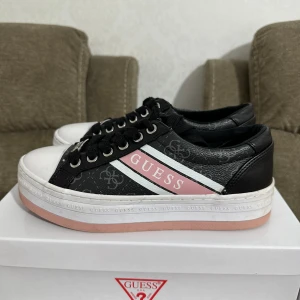 Svarta sneakers från Guess med rosa och vita detaljer - Snygga svarta sneakers från Guess med logomönster, vita tår och rosa sula. Skorna har svarta skosnören, metallfärgade öljetter och en vit rand med rosa GUESS-logga på sidan. Perfekta för en trendig vardagslook.