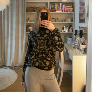 Svart och guldmönstrad tröja från Scotch & Soda - Säljer en snygg svart och guldmönstrad tröja från Scotch & Soda med stort M framtill. Tröjan har lång ärm, ribbade muddar och ett unikt barockinspirerat mönster. Perfekt för dig som vill sticka ut med en cool och lyxig look.