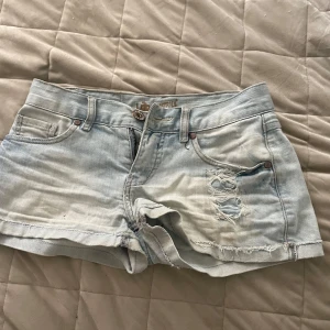 Lågmidjade jeansshorts  - Säljer ett par ljusblå jeansshorts med slitna detaljer och råa kanter. Shortsen har klassisk femficksdesign, bälteshällor och knappgylf. Perfekta för varma dagar och en avslappnad stil. På ena fickan finns det en liten svart fläck! Men arnas i fint sick! Är i storlek 164!