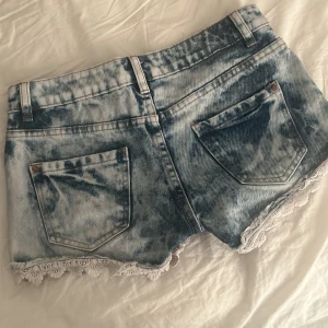 Blå jeansshorts med spetskant från Hotpant - Säljer ett par blå jeansshorts från Hotpant med snygg tvättad effekt och vit spetskant längst ner. Shortsen har normal passform, klassiska fickor och bälteshällor. Perfekta för sommaren! Skulle säga liten i storleken 