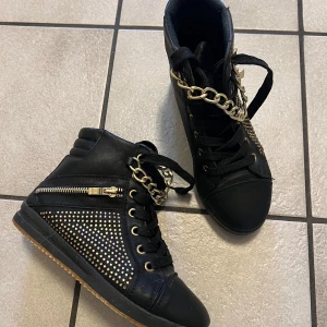 Svarta sneakers med guldkedja och nitar - Säljer ett par svarta sneakers med guldfärgade kedjor och nitar på sidorna. Skorna har snörning, dragkedja på sidan och platt sula. Snygga detaljer som ger en edgy look. Perfekta för dig som vill sticka ut.