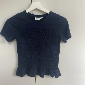 Mörkblå topp med volangkant - Säljer en mörkblå topp med korta ärmar och smockad framsida och baksida. Toppen har en söt volangkant nedtill och rund halsringning. Perfekt för dig som gillar en feminin och trendig stil.