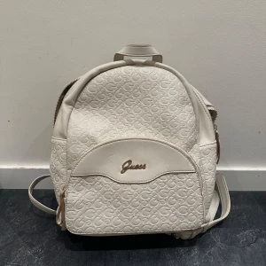 Krämvit ryggsäck från Guess - Säljer en stilren vit ryggsäck från Guess med präglat logomönster och guldfärgad logga framtill. Väskan har flera fack inuti och justerbara axelremmar. Perfekt för dig som vill ha en snygg och praktisk accessoar till vardagen.