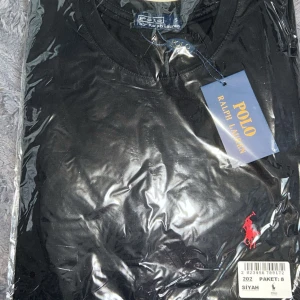 Svart t-shirt från Polo Ralph Lauren - Klassisk svart t-shirt från Polo Ralph Lauren med rund hals och ikoniska röda broderade loggan på bröstet. Perfekt basic-plagg med snygg passform och mjukt bomullsmaterial.
