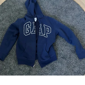 Blå hoodie från GAP med dragkedja - Mörkblå hoodie från GAP med stor logga framtill och dragkedja. Jackan har huva och är fodrad med mjuk teddy på insidan för extra värme. Perfekt för kyliga dagar och enkel att matcha med jeans eller träningsbyxor.