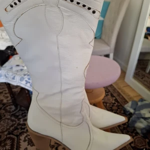 Vita cowboy boots med klack - Säljer ett par vita cowboy boots med spetsig tå och markerad klack. Skorna har dekorativa sömmar och håldetaljer upptill. Perfekta för dig som vill sticka ut med en cool westernstil.