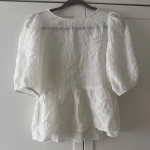 Vit blus med puffärm  - Säljer en luftig vit blus från H&M med puffiga trekvartsärmar och lätt rynk vid axlarna. Blusen har en volang nertill och knäppning bak i nacken. Perfekt för vår och sommar! Genomskinligt tyg