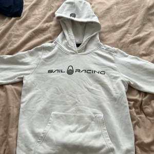 Vit hoodie från Sail Racing - Vit hoodie från Sail Racing med svart logga och text på bröstet. Tröjan har huva, magficka och långa ärmar. Tillverkad i en mjuk bomulls- och polyestermix som känns skön mot huden. Pris kan diskuteras 