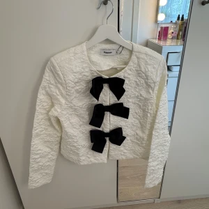 Vit blus/kofta med svarta rosetter från Kappahl - Supersöt vit blus/kofta från Kappahl med strukturmönster och tre stora svarta rosetter framtill. Blusen/koftan har rund halsringning och långa ärmar. Perfekt för dig som vill sticka ut med en unik detalj!