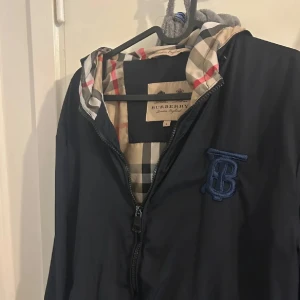 Blå vindjacka från Burberry med klassiskt rutigt foder - Säljer en snygg marinblå vindjacka från Burberry med dragkedja och ikoniskt rutigt foder. Jackan har huva och ett broderat blått Burberry-monogram på bröstet. Perfekt för dig som vill ha en stilren och tidlös jacka.