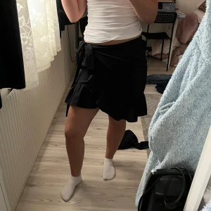 Svart omlottkjol med volang - Säljer en svart omlottkjol med volangkant och knytband i midjan. Kjolen har en medel längd och är perfekt för dig som gillar en enkel men ändå trendig stil. Passar till många olika toppar och är lätt att matcha.