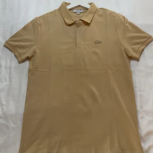 Beige pikétröja från Lacoste - Säljer en klassisk beige pikétröja från Lacoste med korta ärmar och krage. Tröjan har en liten broderad logga på bröstet och knäppning vid halsen. Perfekt för en stilren och avslappnad look.