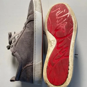 Gråa sneakers från Christian Louboutin - Säljer ett par gråa sneakers från Christian Louboutin med klassisk röd sula och snörning. Skorna har rund tå och är tillverkade i mocka med vita detaljer på sulan. Perfekta för dig som gillar stilrena och exklusiva sneakers. Storlek 42 men passar även 43. 