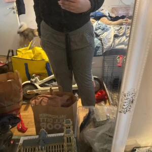 Grå mjukisbyxor med spetsdetalj - Säljer ett par grå mjukisbyxor med svart spetsdetalj upptill och broderad dekoration på ena benet. Byxorna har normal passform och dragsko i midjan för justerbarhet. Perfekta för en chill dag hemma eller när du vill vara extra bekväm.