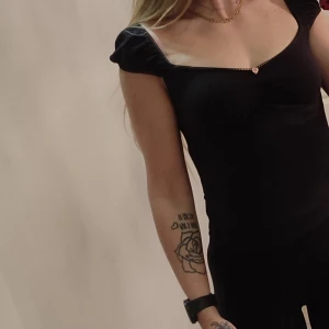 Svart topp med puffärmar - Säljer en svart topp med söt hjärtformad knapp framtill och puffiga korta ärmar. Toppen har en fyrkantig urringning och är figurnära i modellen. Perfekt för att lyfta en enkel outfit.