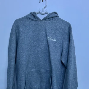 Grå Corteiz hoodie med broderad logga - Grå hoodie i mjuk bomulls- och polyestermix med broderad vit logga på bröstet. Klassisk känguruficka framtill och huva. Perfekt för en avslappnad stil.