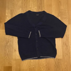 Mörkblå kofta från Zara - Säljer en stilren mörkblå kofta från Zara Man. Koftan har v-ringning, knäppning framtill med fyra knappar och långa ärmar. Perfekt att slänga över en t-shirt eller skjorta för en snygg lager-på-lager-look.