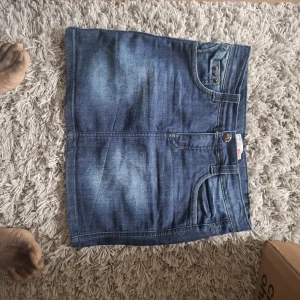 Jeanskjol från Levi's - Säljer en klassisk blå jeanskjol från Levi's med fickor både fram och bak samt knapp- och dragkedjestängning. Kjolen har en rak passform och är i en kort modell, perfekt till sommaren.