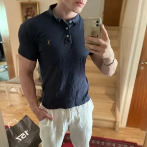 Mörkblå pikétröja från Polo Ralph Lauren - Säljer en mörkblå pikétröja från Polo Ralph Lauren med klassisk krage och knappslå. Tröjan har en liten broderad logga på bröstet och är i slim fit-modell. Perfekt för en stilren och avslappnad look.