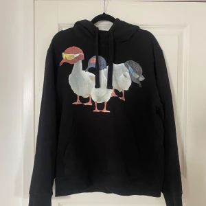 Svart hoodie med ankmotiv från Hugo - Svart hoodie från Hugo med tryck av tre ankor med kepsar på bröstet. Tröjan har huva med dragsko och en stor magficka. Perfekt för dig som gillar streetwear och vill sticka ut med ett unikt motiv.