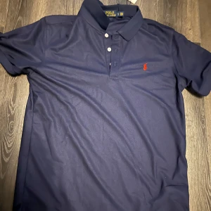 Mörkblå piké t-shirt från Polo Ralph Lauren - Säljer en klassisk mörkblå piké t-shirt från Polo Ralph Lauren med röd broderad logga på bröstet. T-shirten har korta ärmar, krage och tre knappar framtill. Perfekt för en stilren och sportig look.