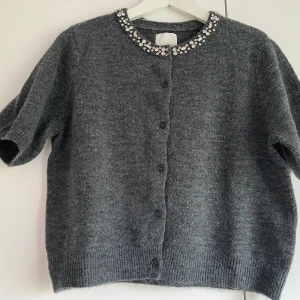Grå kofta med pärldetaljer från H&M - Mysig grå kofta från H&M med korta ärmar och knappar framtill. Kragen är dekorerad med glittrande pärlor och stenar som ger en extra touch. Perfekt att slänga på sig över en topp för en snygg look.