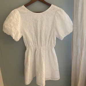 Vit broderad klänning med puffärm från Gina Tricot - Superfin vit klänning från Gina Tricot med broderade blommor, puffärmar och öppen rygg med knyt. Klänningen har en markerad midja och är kort i modellen. Perfekt för sommarens alla tillfällen!