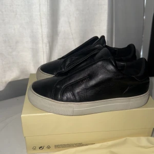 Svarta sneakers från Axel Arigato - Säljer mina skor från Axel Arigato i skinn med vit  sula. Skorna har minimalistisk design utan snörning och diskret logga i guld på sidan som försvunnit lite. Perfekta för en stilren look och kommer och med originalpåse.Annars är skorna helt felfria inga hål ingen skada.Priset kan diskuteras vid snabb affär