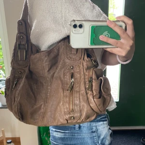 Brun axelväska med flera fack - Säljer en brun axelväska i syntetmaterial med flera dragkedjefack och praktiska sidofickor. Väskan har breda axelband och detaljer i mässingsfärgad metall. Perfekt för dig som vill ha plats för mycket och gillar en avslappnad stil! Kommer med ett extra axelremsband (se sista bild). Verkligen JÄTTESNYGG och passar till ALLT!!💗💗kom med frågor!!
