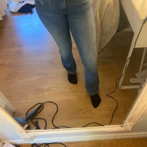 Blå bootcut jeans med mellan midja - Säljer ett par klassiska blå jeans med bootcut och mellan midja. Jeansen har snygga bakfickor med knappdetalj och är i ett mjukt denimtyg. Perfekta till vardags för en avslappnad men trendig look.