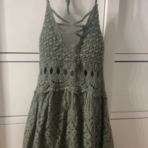 Grön virkad klänning med knyt i ryggen - Supersöt grön klänning med virkad överdel och snygga knytband i ryggen. Klänningen har ärmlös design och mönstrad kjol i spetsliknande material. Perfekt för varma dagar och sommarhäng.