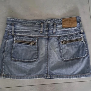 Jeanskjol med dragkedjedetaljer - Säljer en kort jeanskjol i ljusblå denim med två stora fickor och dragkedjor bak. Kjolen har knapp- och dragkedjestängning framtill samt bälteshällor. Perfekt för dig som gillar en avslappnad och trendig stil.