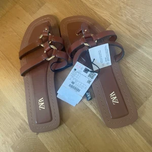 Bruna sandaler från Zara med gulddetaljer - Säljer ett par snygga bruna sandaler från Zara med fyrkantig tå och dekorativa guldfärgade ringar. Remmarna är i brunt syntetmaterial och sulan är platt. Perfekta för sommaren!