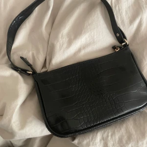 Svart axelväska i croco-mönstrat skinn - Säljer en svart axelväska i croco-mönstrat skinnimitation. Väskan har en enkel design med dragkedja upptill och justerbart axelband. Perfekt storlek för det viktigaste och passar till många olika outfits.