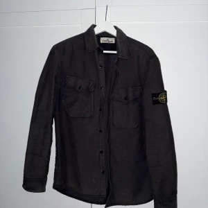 Svart overshirt från Stone Island - Säljer en svart overshirt från Stone Island med klassisk logotyp-patch på vänster ärm. Skjortan har två bröstfickor med knappar och stängs med knappar framtill. Perfekt att bära som lager över en t-shirt. ! Sol blekt !