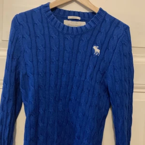 Blå kabelstickad tröja från Abercrombie & Fitch - Säljer en blå kabelstickad tröja från Abercrombie & Fitch med rund halsringning och vit broderad älg-logga på bröstet. Tröjan har lång ärm och är perfekt för dig som gillar klassisk stil med en twist.