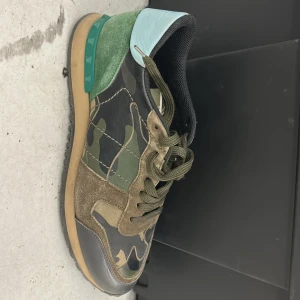 Valentino Garavani Rockrunner sneakers med camouflage - Snygga Valentino Rockrunner sneakers med camouflage-mönster i grönt, svart och beige. Skon har detaljer i mocka, skinn och syntet samt en unik grön häl med nitar och ljusblå bakdel. Klassisk snörning och platt sula.fråga om det är något
