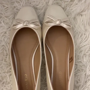 Vita ballerinaskor med rosett - Säljer ett par stilrena vita ballerinaskor från Anna Field med söt rosettdetalj framtill. Skorna har låg klack och är tillverkade i ett syntetmaterial med en lätt glansig finish. Endast använd 1 gång men finns en skavank som inte syns i bilden, skicka ett medelande så får du en bild på det