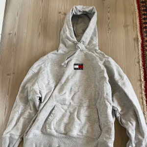Grå hoodie från Tommy Jeans - Säljer en stilren grå hoodie från Tommy Jeans i storlek M. Tröjan har en klassisk huva med dragsko, magficka och en liten logga på bröstet. Perfekt för en avslappnad och trendig look.