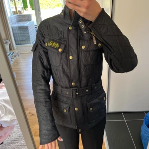 Svart quiltad jacka från Barbour International - Säljer en svart quiltad jacka från Barbour International med guldiga knappar och bälte i midjan. Jackan har hög krage, dragkedja och tryckknappar framtill samt två bröstfickor och två fickor nedtill. Klassisk och stilren modell.