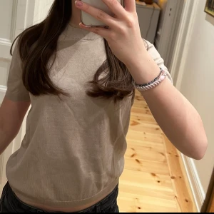 Beige stickad t-shirt från Lindex - Säljer en stilren beige stickad t-shirt från Lindex i mjuk bomull. Toppen har korta ärmar och rund halsringning med ribbad kant. Perfekt för vår och sommar, enkel att matcha till olika outfits.