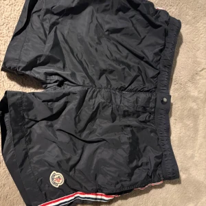 Svarta badshorts från Moncler - Svarta badshorts från Moncler med elastisk midja och knappstängning. Shortsen har Moncler-logga på vänster ben och röd-vit-blå rand längs benslutet NFC TAG FUNKAR. Perfekta för beachen och brudarna🤩😉! (Pris kan diskuteras)