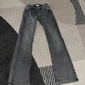 Gråa låg midjade jeans från Gina nytt skick - Grå jeansbyxor från Gina Tricot med raka ben och snygga bakfickor med lock och knapp. Byxorna har normal passform och klassisk knappstängning framtill. Perfekta för en avslappnad och stilren look. Köparen står för frakten