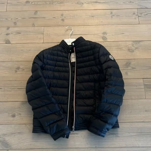 Svart pufferjacka från Moncler - Säljer en svart pufferjacka från Moncler med klassisk quiltad design och rund krage. Jackan har dragkedja framtill och Moncler-logga på ärmen. Perfekt för kyliga dagar och riktigt snygg till vardags. Nypris 6000kr storlek 14 years